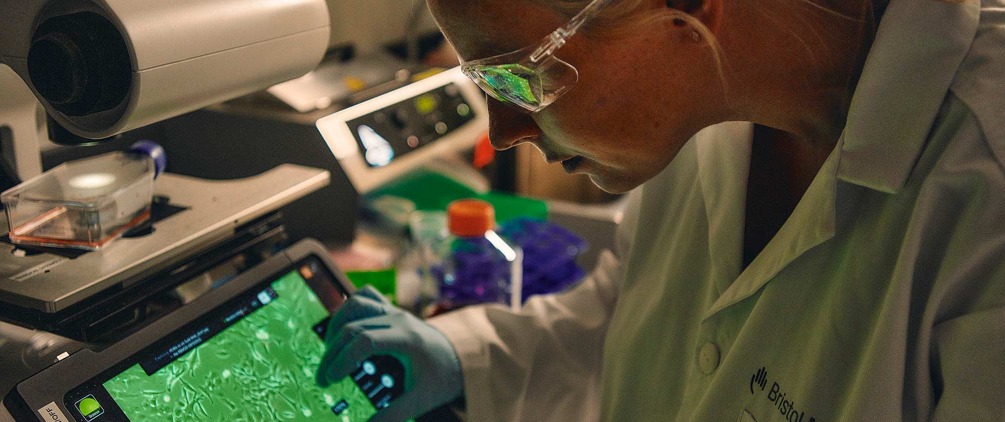 A researcher wearing a Bristol Myers Squibb lab coat and safety glasses uses her fingers to zoom in on a tablet screen.