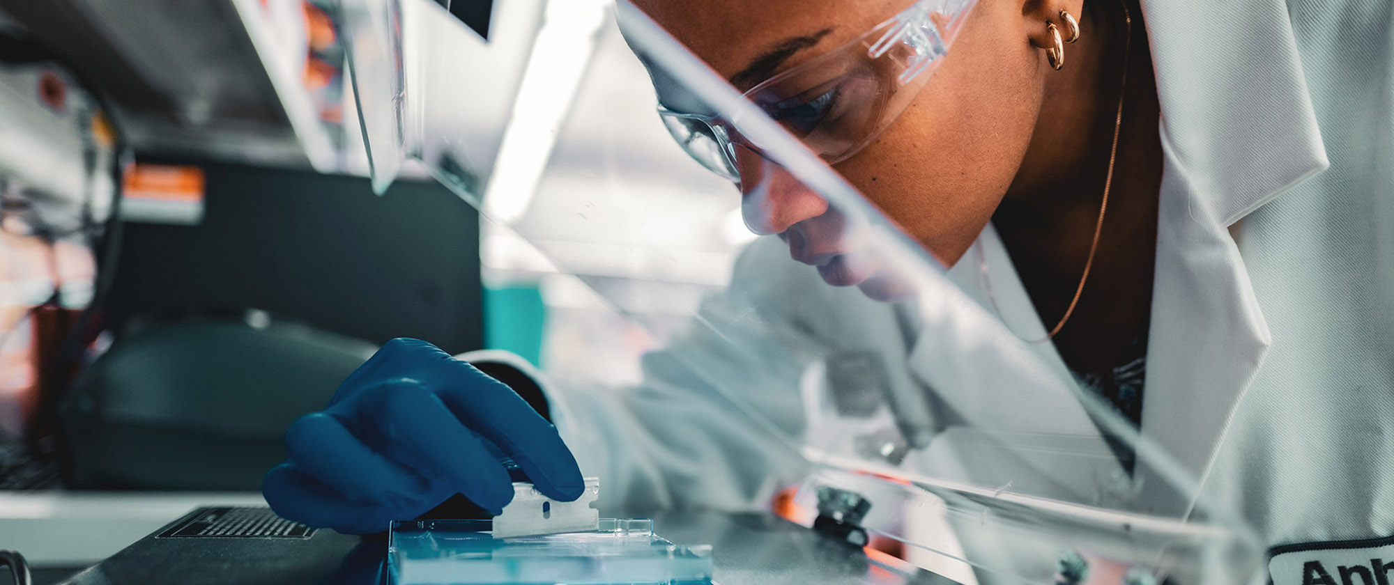 Female scientist wearing protective safety glasses wearing a white lab coat and blue gloves, uses a razor blade on a streak plate. 